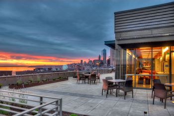 A rooftop deckpatio with chairs and a table overlooking a city skyline at sunset.